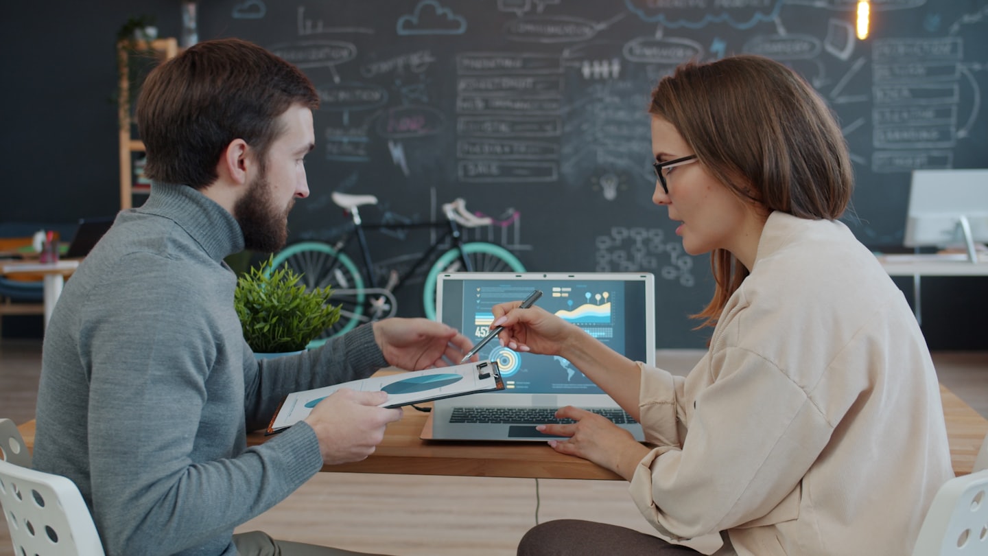 Two business people discuss data on a laptop and document.